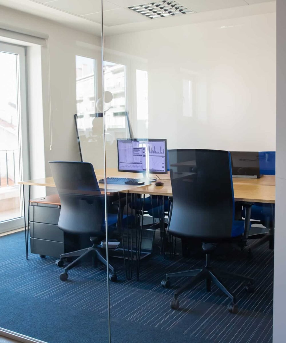 Empty boardroom behind glass wall. Meeting room with conference table, shared desk for team and workplaces. Trading graphs on monitor. Office interior or commercial real estate concept